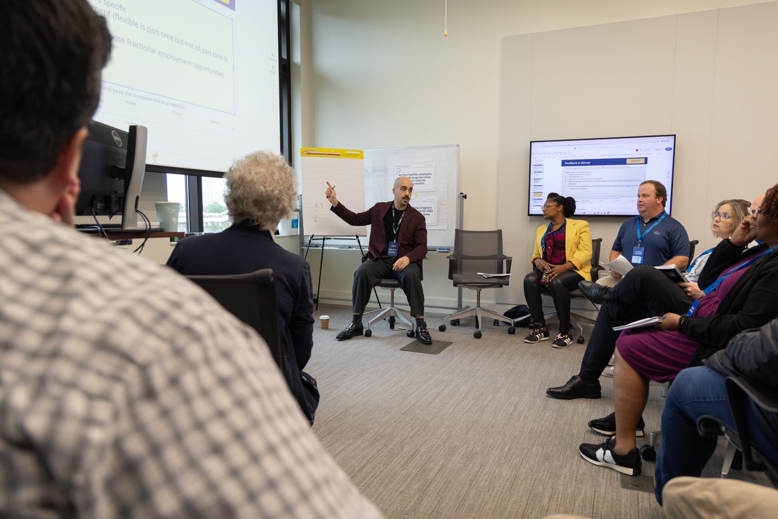 A facilitator leads a small working group seated in a circle, with draft policy feedback on the screens behind them